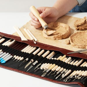 Puede incluir: Un completo juego de herramientas de cerámica con varias herramientas de escultura, incluyendo herramientas con mango de madera, cuchillas metálicas y pinceles. Las herramientas se guardan en un estuche enrollable de cuero marrón. Una mano está usando una herramienta para esculpir arcilla.