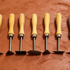 May include: A set of seven clay sculpting tools arranged in a row, each featuring a smooth, light-colored wooden handle, a silver metal ferrule, and a unique metal blade shape, including triangular, square, and rounded tips for detailed work.