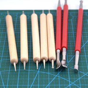 May include: A collection of eight clay sculpting tools. Six tools have natural wood handles and metal tips. The other two tools have red handles and metal tips. The tools are arranged on a green cutting mat.