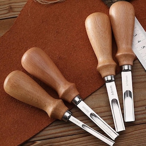 Può includere: Quattro strumenti per la lavorazione della pelle con manici in legno e lame in acciaio inossidabile. Le lame sono etichettate A4mm, A6mm, A8mm e A10mm. Gli strumenti sono disposti su una superficie in pelle marrone.