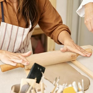 Puede incluir: Un rodillo de madera de color claro utilizado para aplanar una placa de arcilla gris. El rodillo presenta un cuerpo cilíndrico con mangos cónicos en cada extremo, colocado horizontalmente sobre la superficie de la arcilla al trabajar.