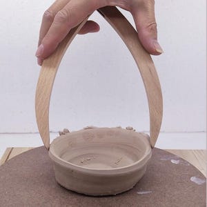 May include: A light brown clay bowl with a circular base and a smooth interior, featuring two tall, curved wooden handles attached to opposite sides of the rim. The bowl has a natural, unglazed finish with visible texture and small clay fragments.