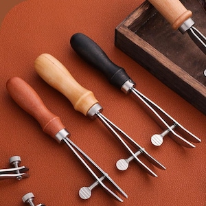 May include: Close-up of leatherworking tools with wooden and black handles. The tools have a silver metal construction with a screw adjustment. The handles are in various colors, including light brown, dark brown, and black. The tools are arranged on a brown surface.