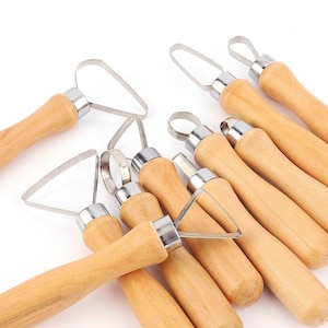 May include: A collection of pottery tools with wooden handles and stainless steel blades. The tools have various blade shapes, including loops and triangles, and are designed for sculpting and shaping clay. The handles are light brown.
