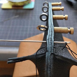 Könnte beinhalten: Nahaufnahme eines Lederriemens mit Metallringen und Messing-Schrauben. Eine Nadel und Faden werden verwendet, um den Riemen zusammenzunähen.