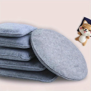 Puede incluir: Una pila de inserciones ovaladas de fieltro azul claro para camas de mascotas. Las inserciones están cuidadosamente apiladas, con un borde cosido visible. Un pequeño juguete de peluche de perro Shiba Inu descansa sobre una almohada azul en el fondo.
