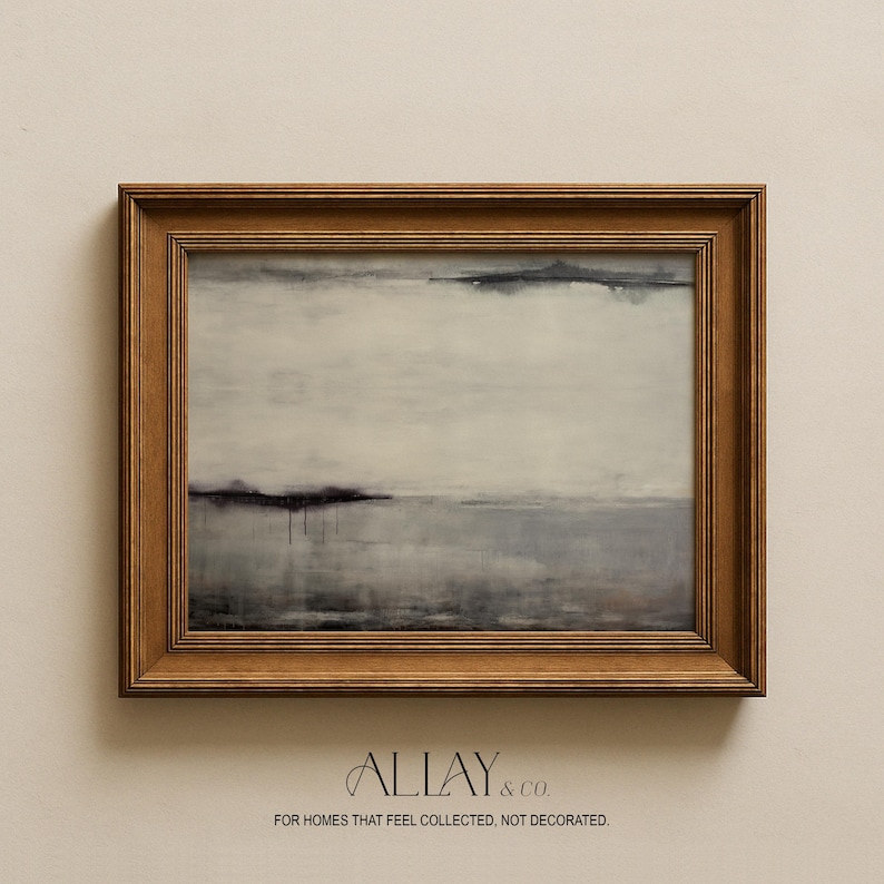 Puede incluir: Una pintura de paisaje rectangular en un marco de madera oscura con bandas horizontales abstractas de blanco, gris y negro con sutiles gotas de pintura. Debajo del marco, el texto dice: ALLAY & CO. FOR HOMES THAT FEEL COLLECTED, NOT DECORATED.