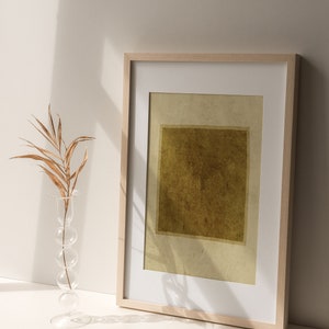 May include: A rectangular framed print featuring a dark olive green square centered on a light beige background, housed in a light wood frame with a white mat. Beside the frame stands a clear glass vase with a stacked sphere design holding a dried leaf.
