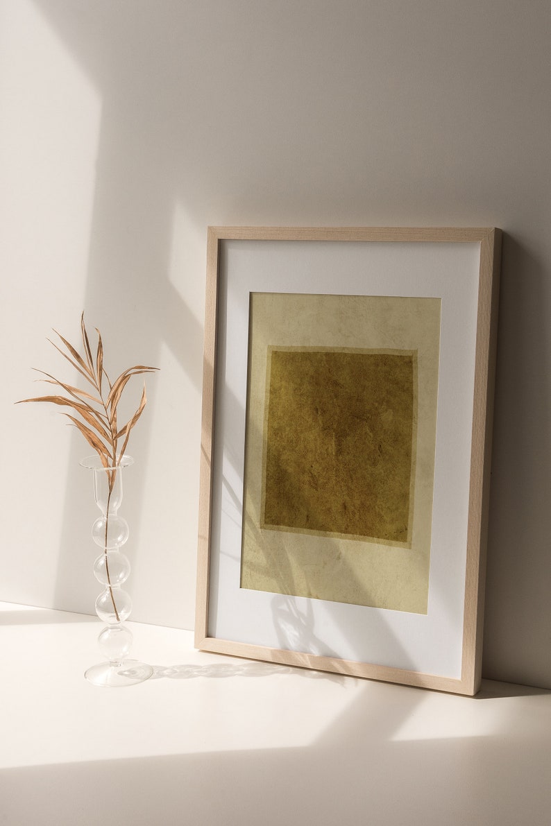 May include: A rectangular framed print featuring a dark olive green square centered on a light beige background, housed in a light wood frame with a white mat. Beside the frame stands a clear glass vase with a stacked sphere design holding a dried leaf.
