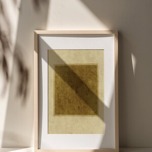 May include: A rectangular wall art print featuring a dark olive-brown square centered on a light beige background, enclosed within a light-toned wooden frame with a white mat border, displaying a textured appearance and soft shadow overlays.