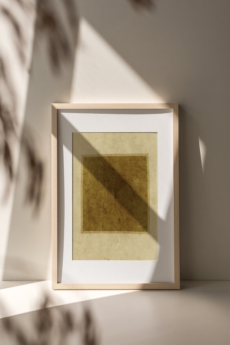 May include: A rectangular wall art print featuring a dark olive-brown square centered on a light beige background, enclosed within a light-toned wooden frame with a white mat border, displaying a textured appearance and soft shadow overlays.
