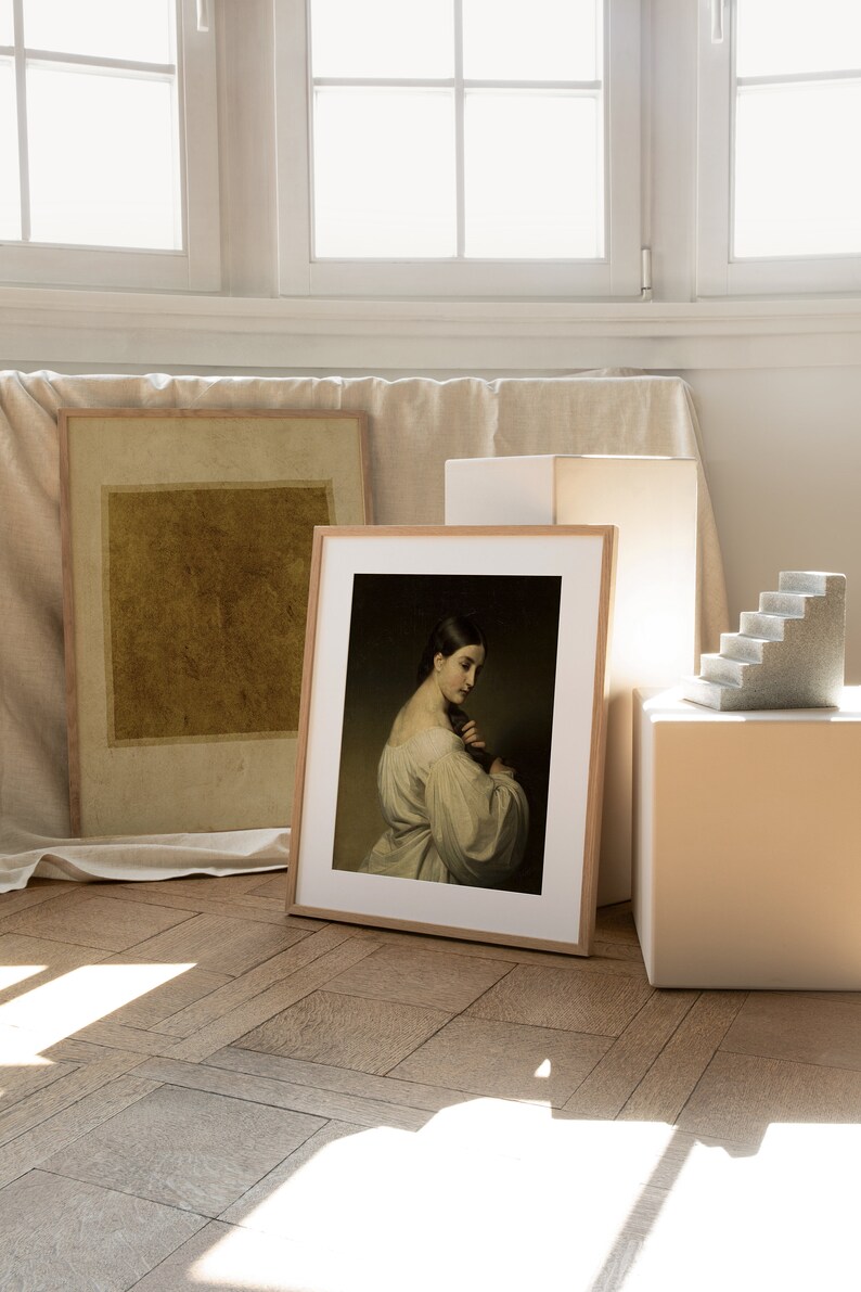 May include: A framed art print featuring a portrait of a person with dark hair in a light-colored garment, positioned next to a second framed print with a solid mustard-yellow square and a small gray stone staircase sculpture on a white block.