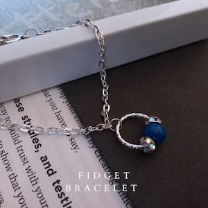 Op de afbeelding: Zilveren kettingarmband met een blauwe kraal en heldere kristallen accenten. De armband heeft een kleine ringbedel met een textuurontwerp. De tekst "FIDGET BRACELET" is zichtbaar op de achtergrond.
