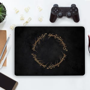 May include: A rectangular black laptop skin featuring a circular gold Elvish script design in the centre. The surface has a subtle textured appearance, and the gold lettering forms a ring shape, mimicking the inscription from The Lord of the Rings.