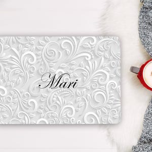 May include: A rectangular laptop skin featuring a white and light grey embossed floral scroll pattern with the name Mari printed in black script in the centre. The design includes intricate swirling vines and leaves across the entire surface.