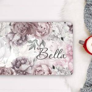Puede incluir: Una funda rectangular para portátil con un estampado floral de peonías en rosa suave, malva y blanco con hojas verdes. El nombre Bella está impreso en una caligrafía cursiva negra en el centro del diseño floral sobre un fondo blanco.