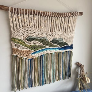 May include: A beige, green and blue macrame wall hanging with wooden beads and a natural wood dowel at the top. The hanging has a fringe at the bottom.