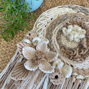 May include: A beige and white macrame wall hanging with a floral design. The hanging features a large flower with a fluffy centre and smaller flowers around it. The macrame is made with different textures and thicknesses of rope.