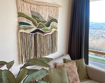 May include: A macrame wall hanging with a landscape design featuring green, brown, and beige yarn. The hanging is attached to a wooden branch and has fringe at the bottom.