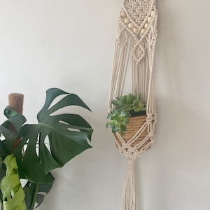 May include: A macrame plant hanger with a wooden dowel and white beads. The hanger is holding a small potted succulent plant.