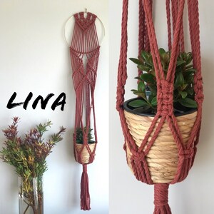 May include: A macrame plant hanger in a terracotta colour, featuring a geometric pattern and a hanging pot. The hanger is labelled "LINA" and is holding a small potted plant.