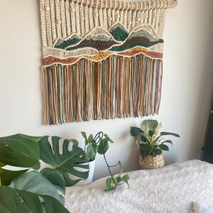 May include: A macrame wall hanging with a mountain landscape design. The hanging features a natural wood dowel, beige, brown, green, and orange yarn, and wooden beads.