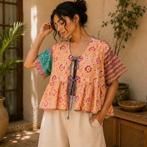 Könnte beinhalten: Ein kurzärmeliges Oberteil im Schößchen-Stil mit einem leuchtend orange-rosa Rautenmuster, einem V-Ausschnitt mit dünnen Bindebändern und kontrastierenden, gemusterten Ärmeln in Petrol und Violett, kombiniert mit einer weiten, hellbeigen Hose.