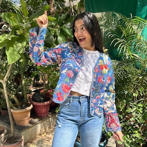 May include: A woman wearing a blue floral patterned jacket with a white shirt and blue jeans. She is standing in a garden setting with plants and potted plants.