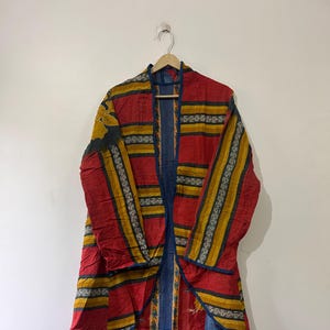 Puede incluir: Una chaqueta larga y abierta en rojo, amarillo y azul. La chaqueta presenta rayas horizontales amarillas y azules sobre un fondo rojo. La parte inferior de la chaqueta tiene un estampado floral con flores amarillas y azules. La chaqueta está colgada en una percha de madera.