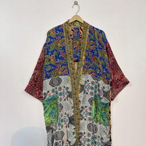 Op de afbeelding: Een kleurrijke kimono-jas met een paisley-patroon in blauw, rood, groen en wit. De jas hangt aan een houten hanger tegen een witte muur. De kimono heeft lange mouwen en een vloeiend silhouet.