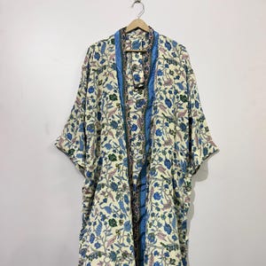 Könnte beinhalten: Lange, offen geschnittene Kimono-Robe mit floralem Muster in Blau-, Grün- und Rosatönen auf cremefarbenem Hintergrund. Die Robe hat eine blaue Bordüre an den Rändern und hängt an einem Holzbügel.