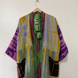 Pode incluir: Um quimono tie-dye em tons de roxo, rosa, amarelo e verde pendurado em um cabide de madeira. O quimono apresenta um design de patchwork com dois bolsos frontais e uma gravata combinando. As mangas são largas e o tecido parece leve.