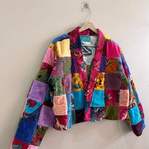 May include: A cropped patchwork jacket with a vibrant mix of colors and patterns. The jacket features a variety of velvet squares in shades of pink, purple, blue, and green, creating a unique and eye-catching design. The jacket is hanging on a wooden hanger.
