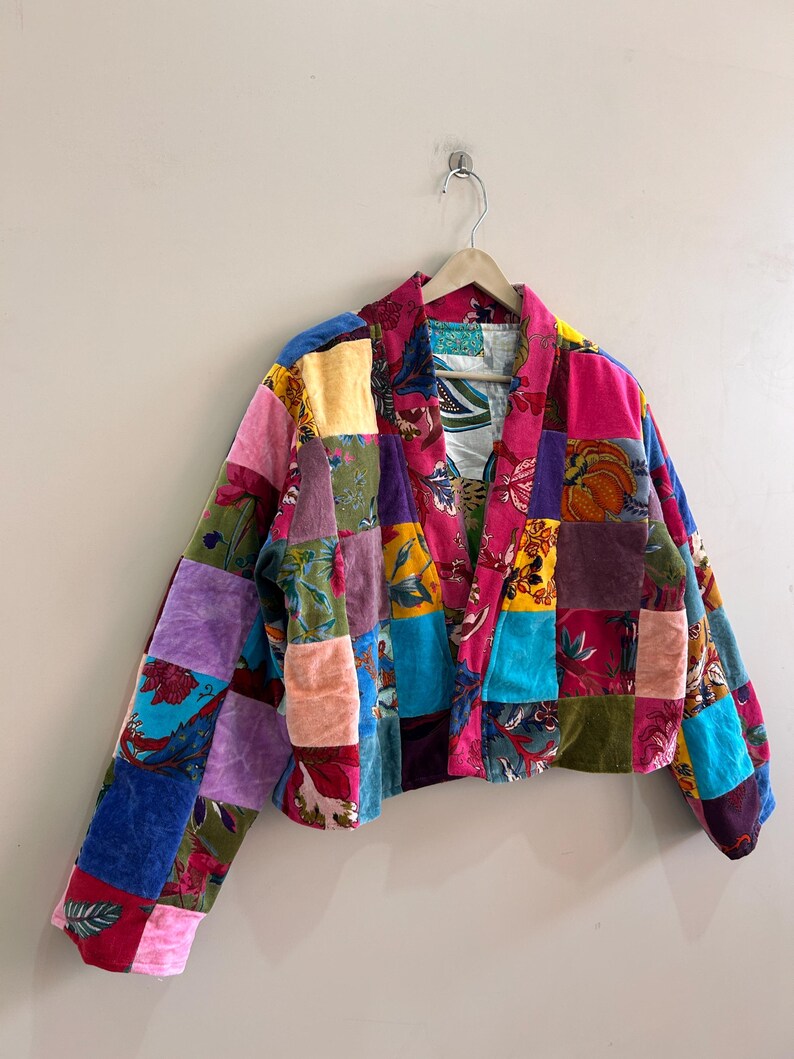 May include: A cropped patchwork jacket with a vibrant mix of colors and patterns. The jacket features a variety of velvet squares in shades of pink, purple, blue, and green, creating a unique and eye-catching design. The jacket is hanging on a wooden hanger.