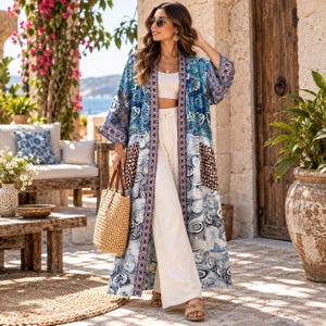 May include: A long, open-front kimono featuring a vibrant blue, white, and brown paisley and floral patchwork pattern. The garment has wide sleeves and a decorative border along the front edges. It is worn over a white crop top and wide-leg pants.