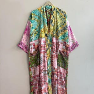 Può includere: Un kimono patchwork colorato con una varietà di motivi e colori, tra cui rosa, verde, giallo e blu. L'abito ha maniche lunghe e due tasche. È appeso a una gruccia di metallo.
