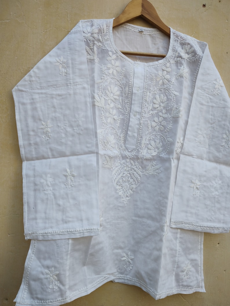 May include: White cotton tunic top with intricate white embroidery featuring floral designs. The top has a round neckline and long sleeves.