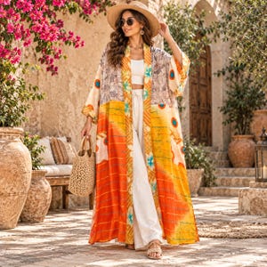 May include: A long, open-front kimono featuring a vibrant pattern of orange, yellow, and teal floral motifs. The garment has wide sleeves and a floor-length hem, worn over a white crop top and white wide-leg trousers, paired with a straw tote bag.