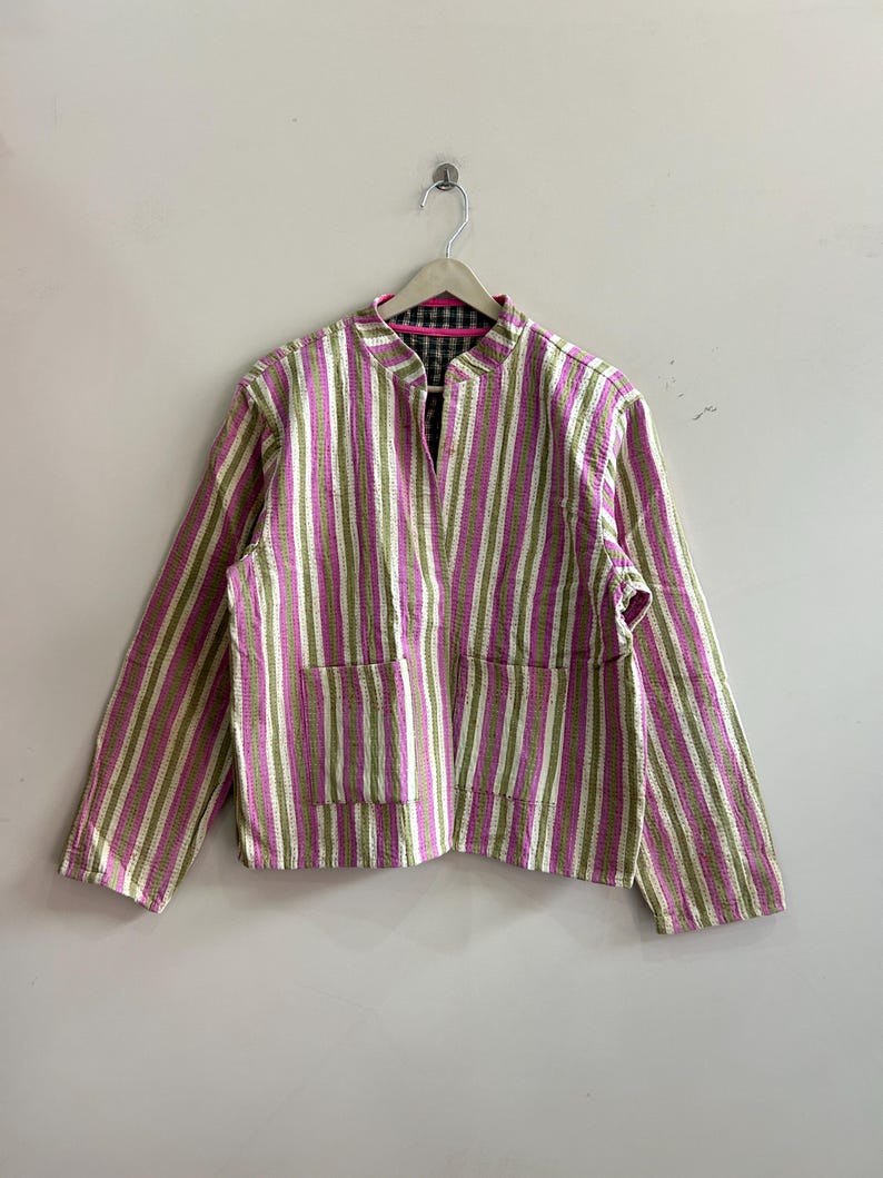 May include: A pink, green, and white striped jacket with a mandarin collar and two front pockets. The jacket is hanging on a wooden hanger against a white wall. The interior lining is a black and green plaid.