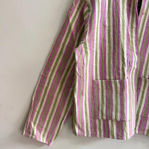 May include: A striped jacket in shades of pink, green, and white. The jacket features a collared neckline, long sleeves, and two front pockets. The fabric has a textured appearance with vertical stripes.