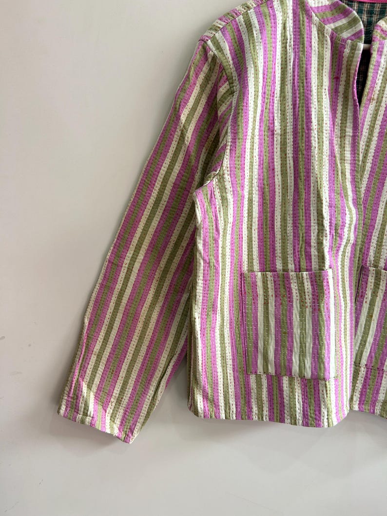 May include: A striped jacket in shades of pink, green, and white. The jacket features a collared neckline, long sleeves, and two front pockets. The fabric has a textured appearance with vertical stripes.
