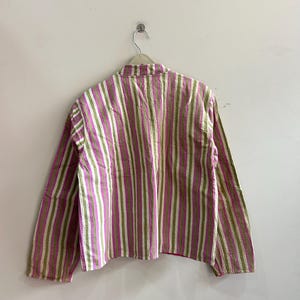 May include: A long-sleeved shirt with vertical stripes in shades of pink, green, and white. The shirt is hanging on a hanger against a plain white wall. The fabric appears to have a textured, quilted appearance.