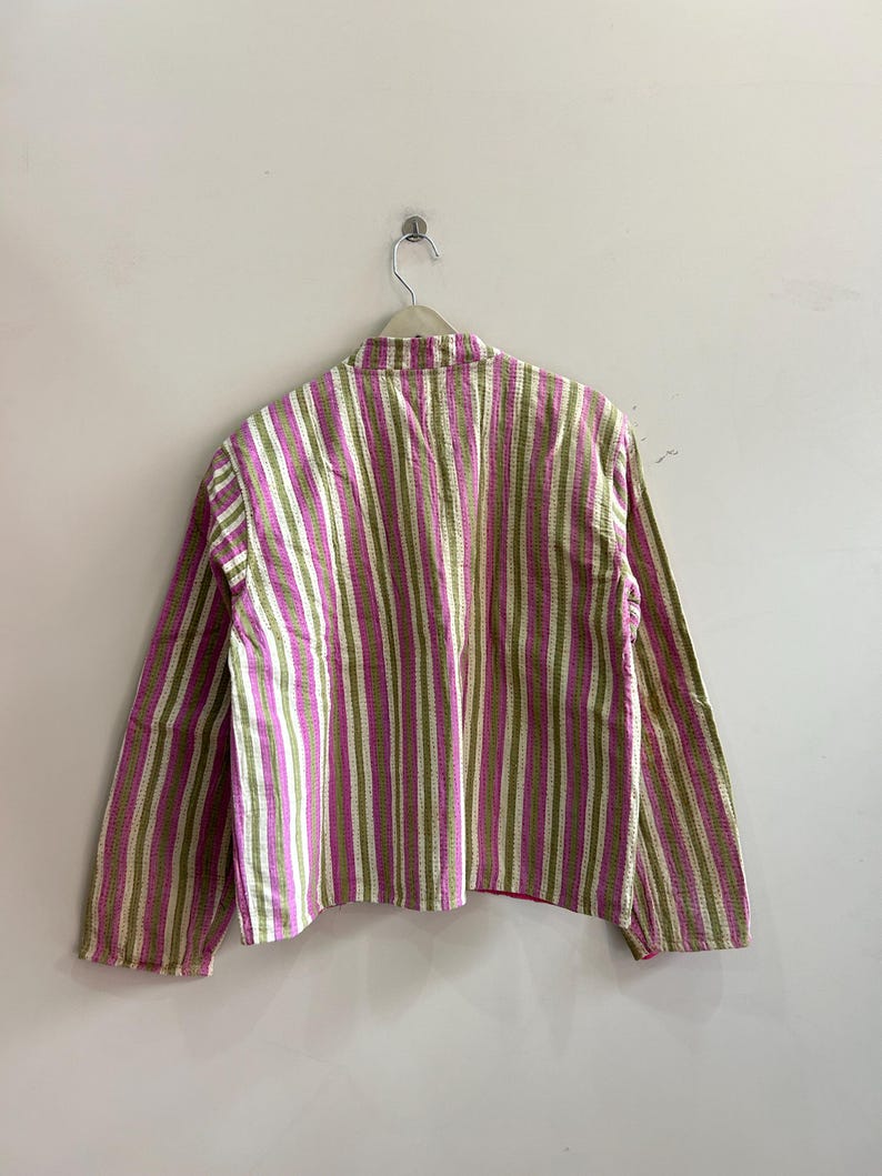 May include: A long-sleeved shirt with vertical stripes in shades of pink, green, and white. The shirt is hanging on a hanger against a plain white wall. The fabric appears to have a textured, quilted appearance.