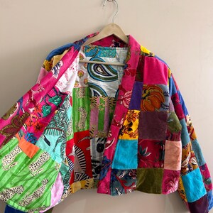 May include: A patchwork jacket with a vibrant mix of colors and patterns, including floral, paisley, and animal prints. The jacket is made of various fabrics and textures, creating a unique and eye-catching design. It hangs on a wooden hanger.