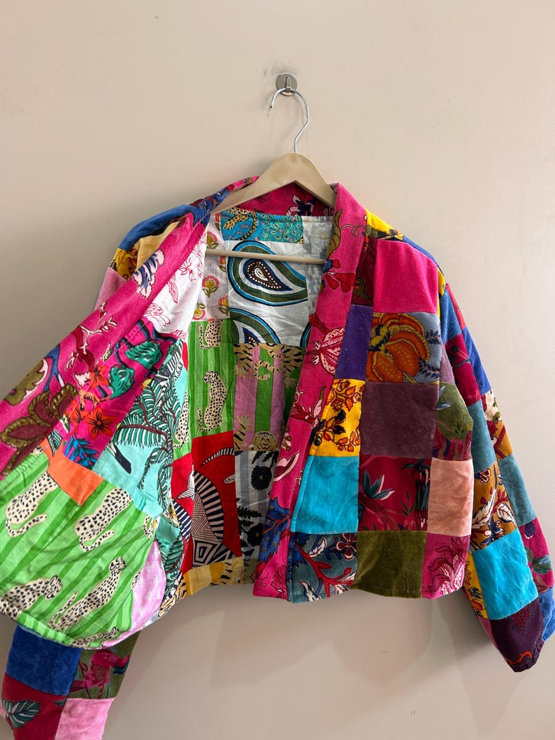 May include: A patchwork jacket with a vibrant mix of colors and patterns, including floral, paisley, and animal prints. The jacket is made of various fabrics and textures, creating a unique and eye-catching design. It hangs on a wooden hanger.