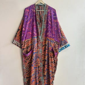 Könnte beinhalten: Ein langärmeliger Kimono-Bademantel mit einem lebendigen Patchwork aus violetten, orangefarbenen und blauen Paisley-Mustern. Das Kleidungsstück hat eine offene Vorderseite, einen breiten Kragen und zwei Vordertaschen mit geometrischen Kontrastborten.
