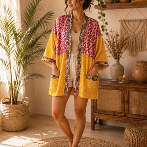 Könnte beinhalten: Eine Jacke im Kimono-Stil mit einem Color-Block-Design, bestehend aus einem leuchtend gelben Unterteil mit weißen floralen Linienmustern und einem Oberteil mit pink-weißem Muster, abgerundet durch eine dekorative florale Bordüre.
