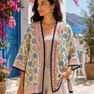 May include: A cream-coloured open-front kimono featuring a vibrant geometric pattern in blue, yellow, and orange. The garment includes intricate embroidery, small reflective mirror accents along the front placket, and dark navy blue trim.