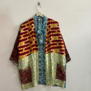 May include: A kimono-style jacket featuring a patchwork design with various patterns and colors. The upper section has red and yellow horizontal stripes, while the lower part displays light green paisley and floral prints with two front pockets.