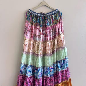 Puede incluir: Una falda larga escalonada con un diseño de patchwork. La falda presenta una variedad de colores, incluyendo rosa, azul, verde y naranja, con estampados de cachemira y florales. La falda está colgada en una percha de madera contra una pared blanca.